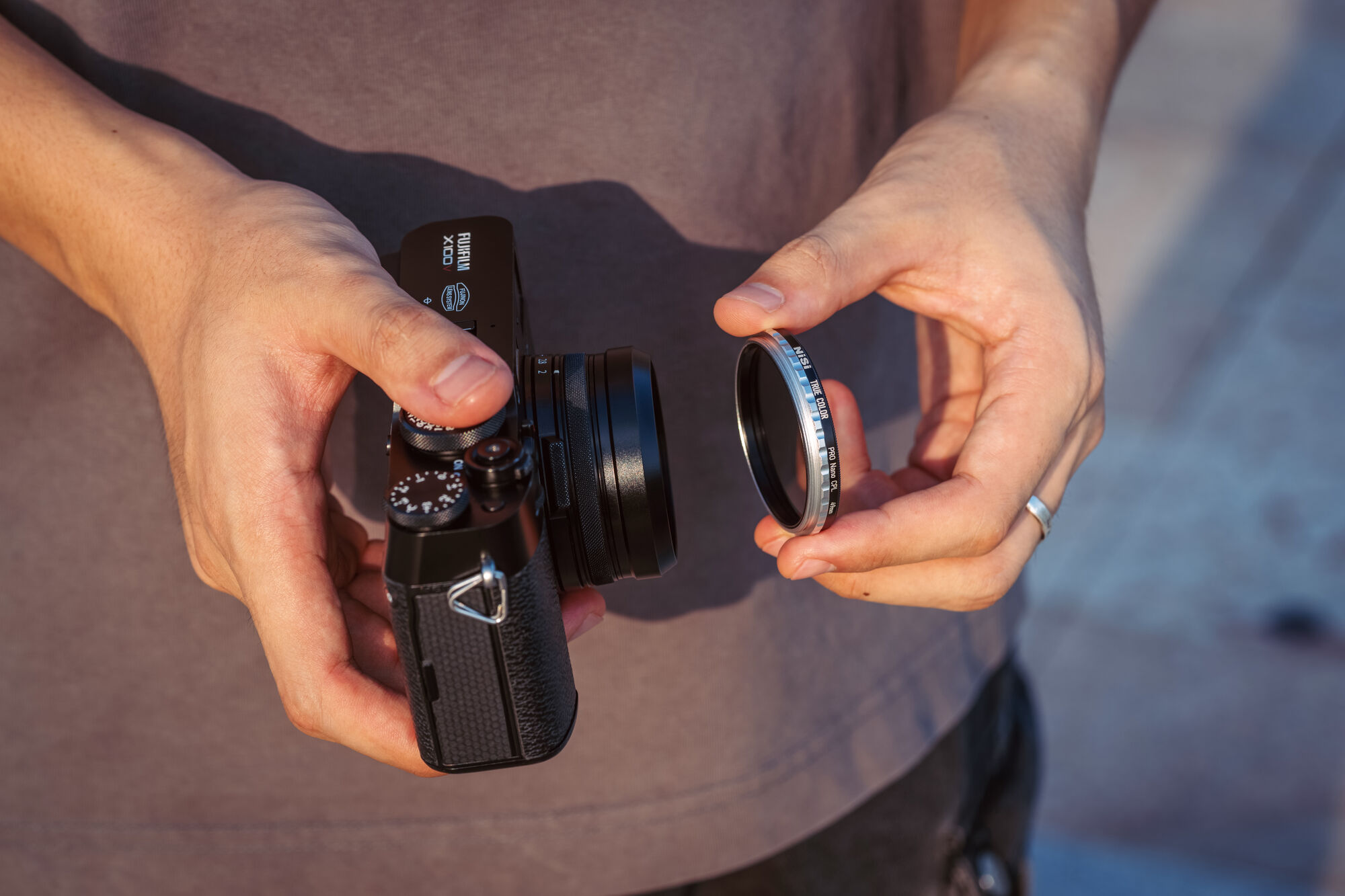 A black Fujifilm X100 held in one hand with the NiSi NC UV Filter II held in the other, about to be threaded on